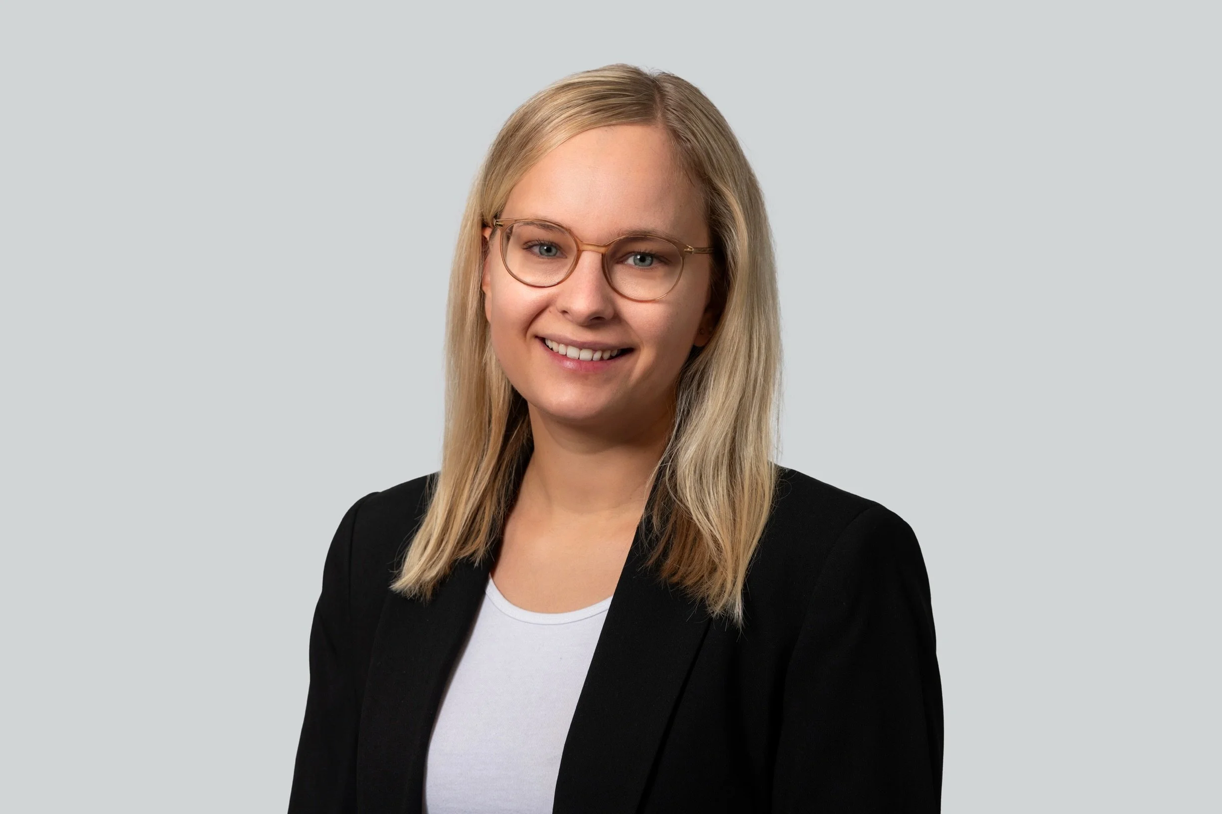 Porträt einer jungen Frau mit blonden Haaren, Brille, im schwarzen Blazer und weißem T-Shirt vor grauem Hintergrund.