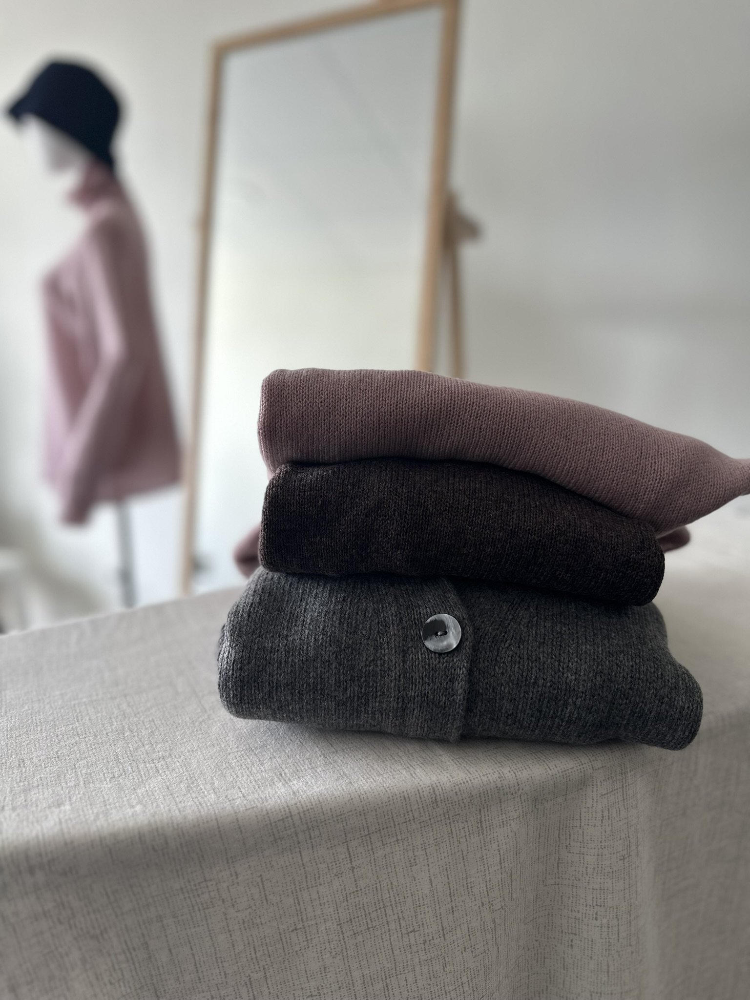 Folded sweaters in gray, black, and pink stacked on a table with a blurred background of a mirror and a mannequin wearing a pink coat and a black hat.