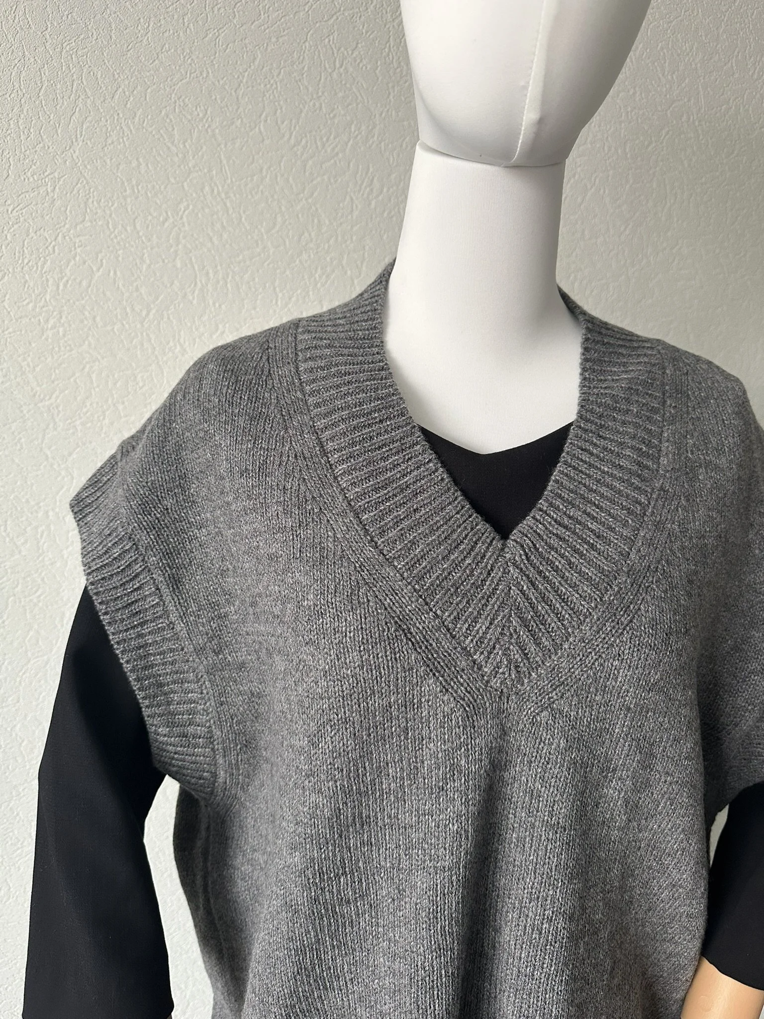 A mannequin wearing a gray sleeveless knit sweater over a black long-sleeve shirt, against a textured white wall.