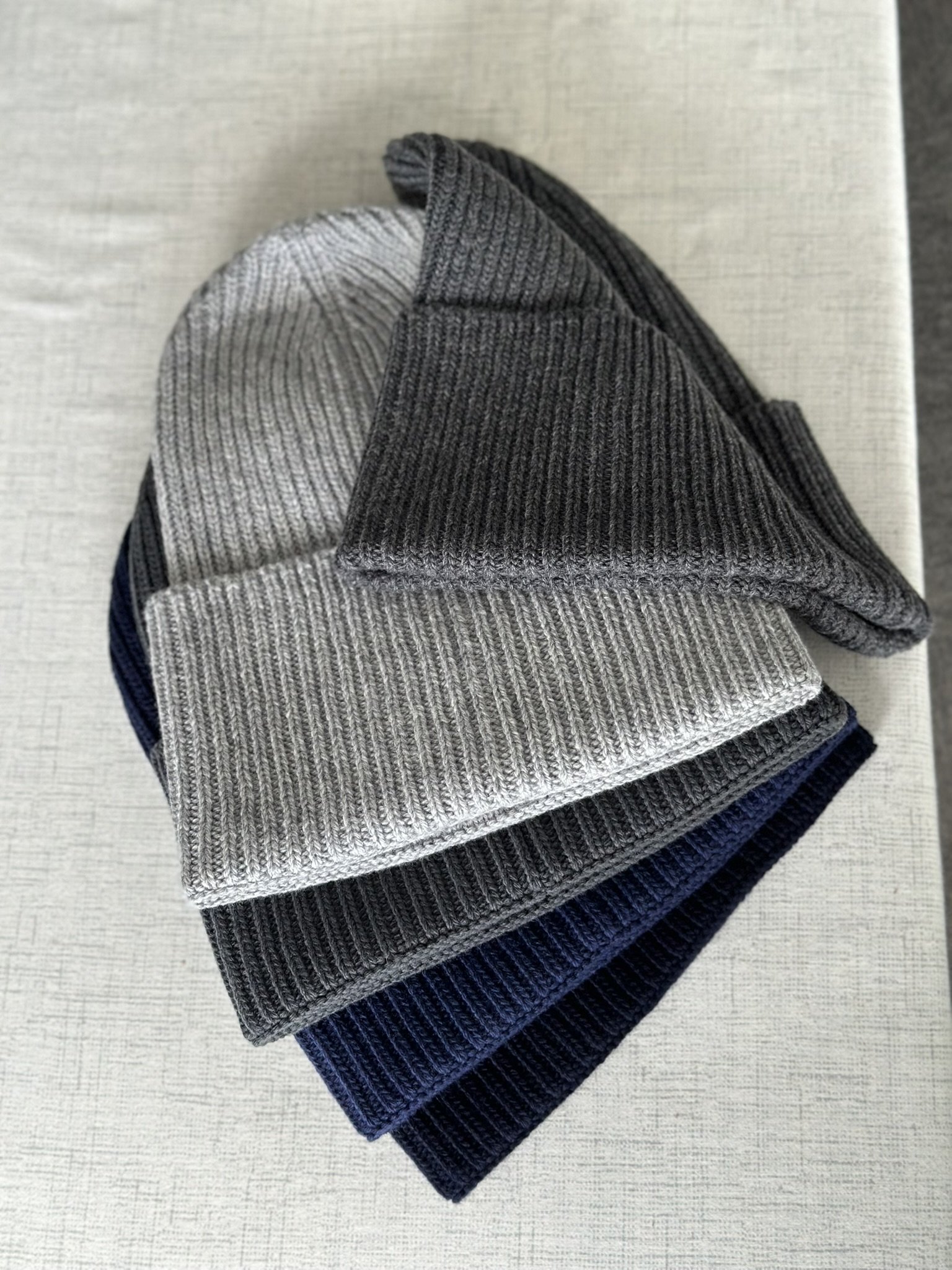 A group of gray, dark gray, navy blue, and black knit beanies stacked on a beige surface.
