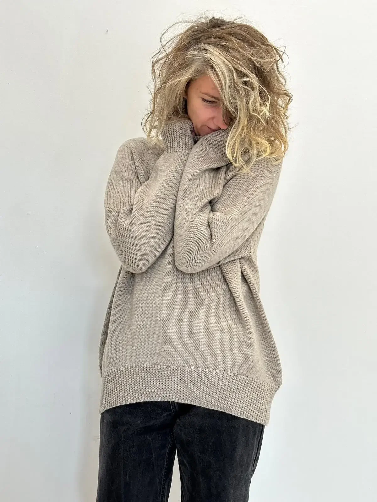 A woman with curly blonde hair, wearing a beige sweater, holding her collar with her hands, standing against a plain white wall, looking down and slightly to the side.