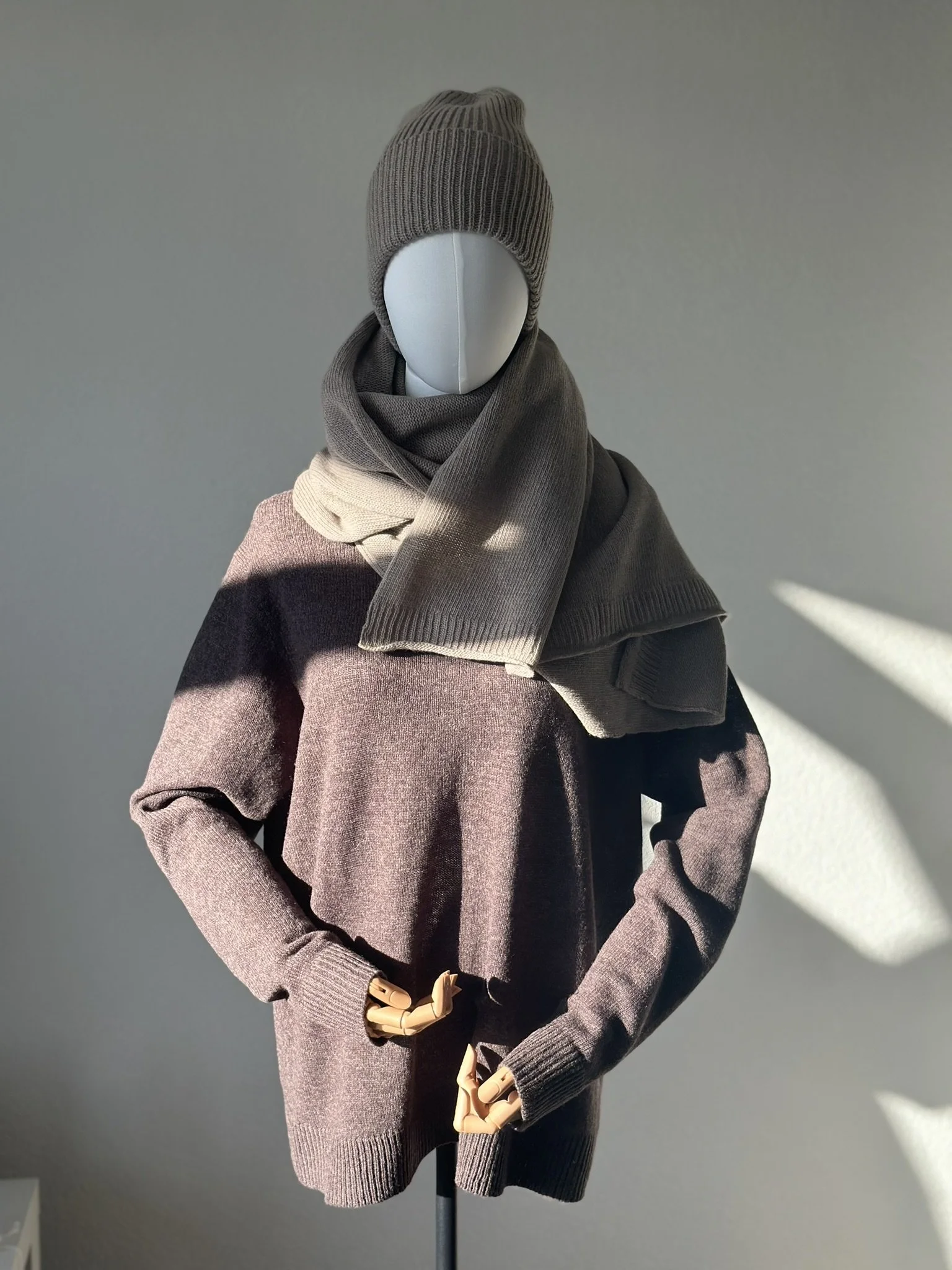 A mannequin dressed in a gray beanie, scarf, and sweater with long sleeves, standing against a plain wall with sunlight casting shadows.