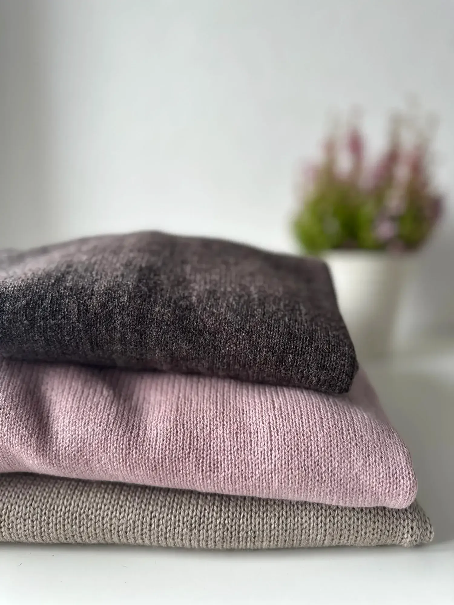 Stack of three folded sweaters in gray, pink, and beige, with a blurred pink flower bouquet in the background.