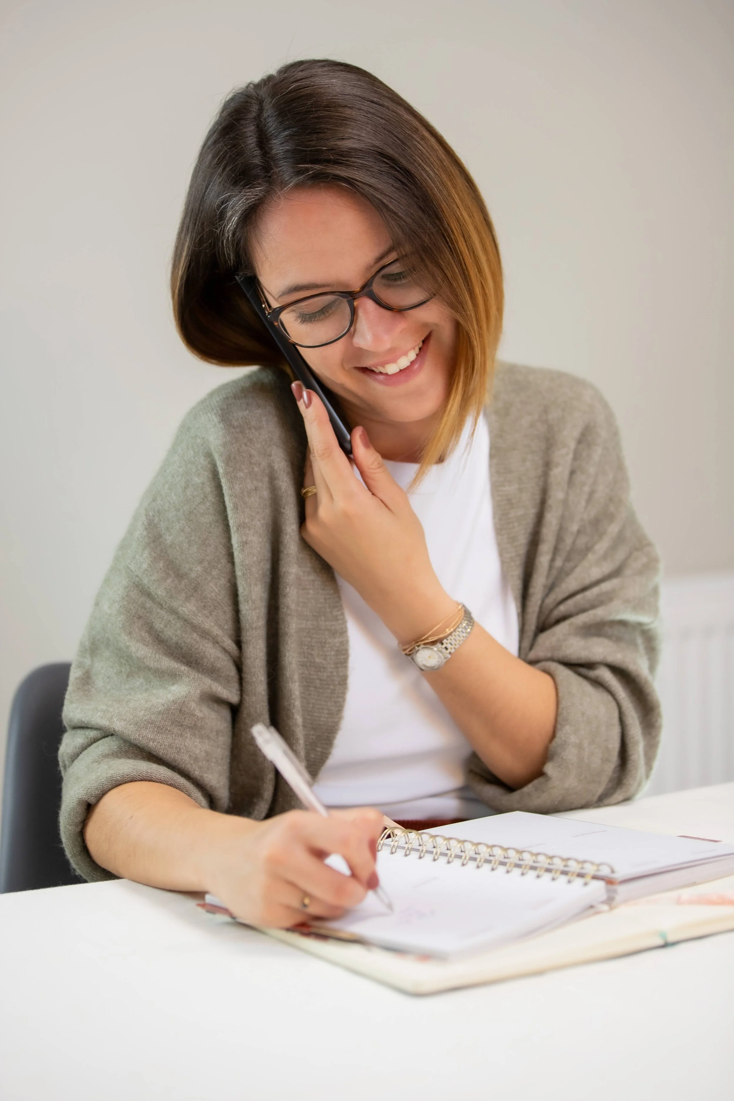 Femme souriante avec lunettes, portant un chandail beige, parlant au téléphone tout en écrivant dans un carnet blanc.