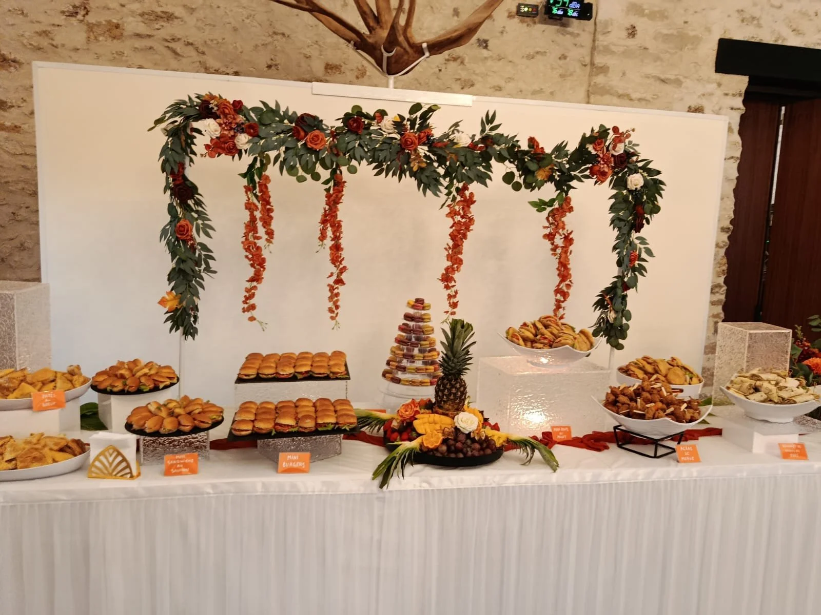 Un buffet de desserts et amuse-bouches décoré avec une arche de feuillage et de fleurs, avec diverses douceurs telles que petits sandwiches, macarons, fruits et autres pâtisseries, dans un cadre intérieur