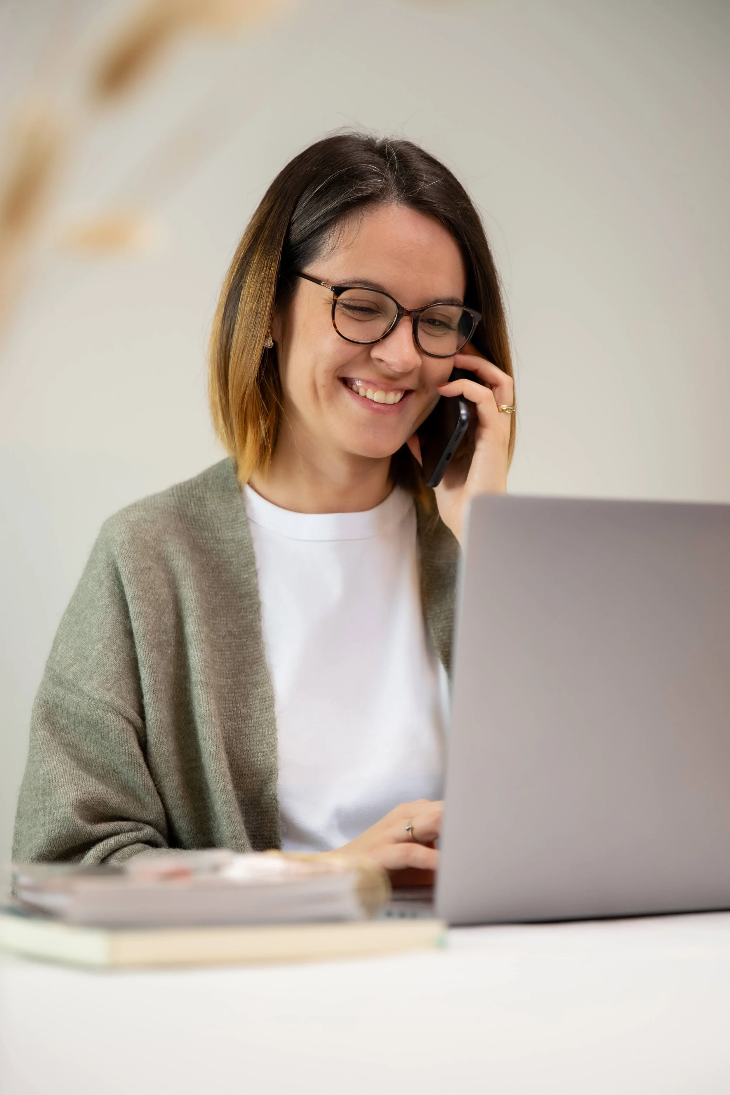 Femme souriante avec des lunettes, parlant au téléphone, travaillant sur un ordinateur portable dans un bureau.