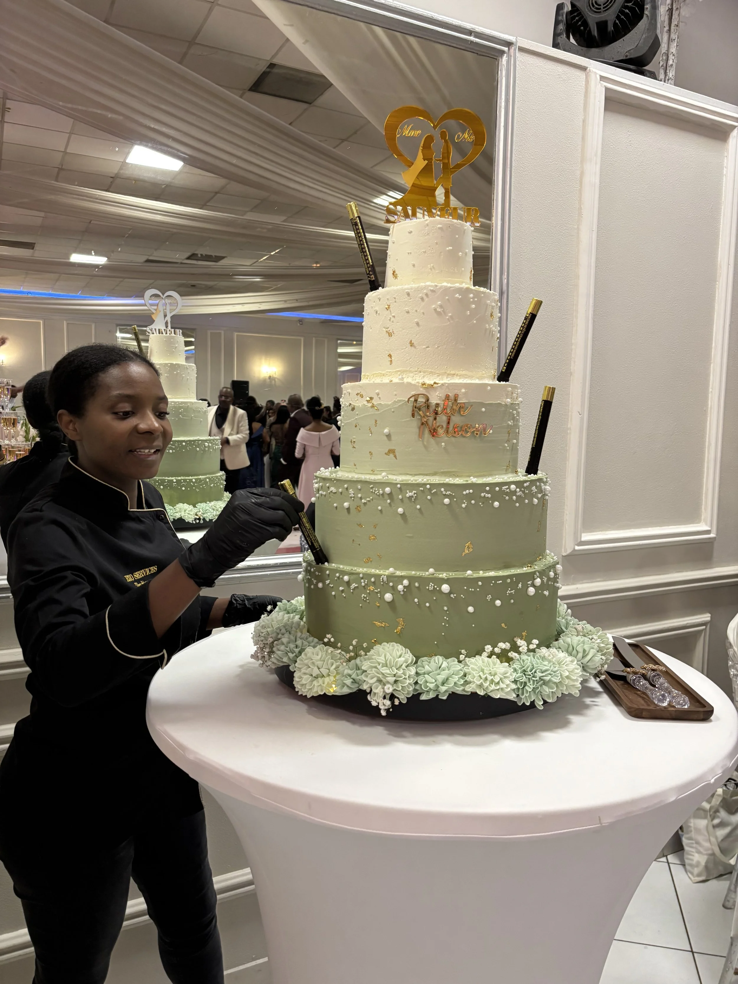 Un gâteau de mariage à plusieurs niveaux décoré de détails blancs et dorés, avec une sculpture en haut représentant un couple dans un cœur. Une femme en noir est en train de décorer le gâteau lors d'une célébration au sein d'une salle élégante remplie de personnes.