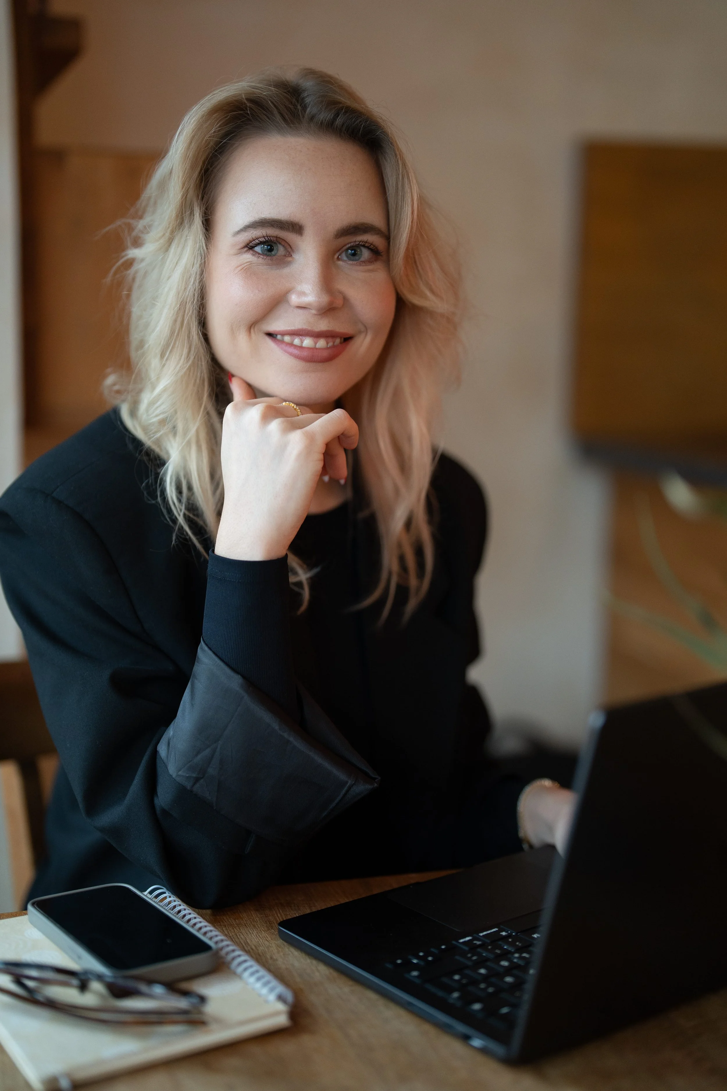 A young woman with blonde, wavy hair, sitting at a desk with a laptop, smartphone, notebook, and glasses, smiling at the camera.