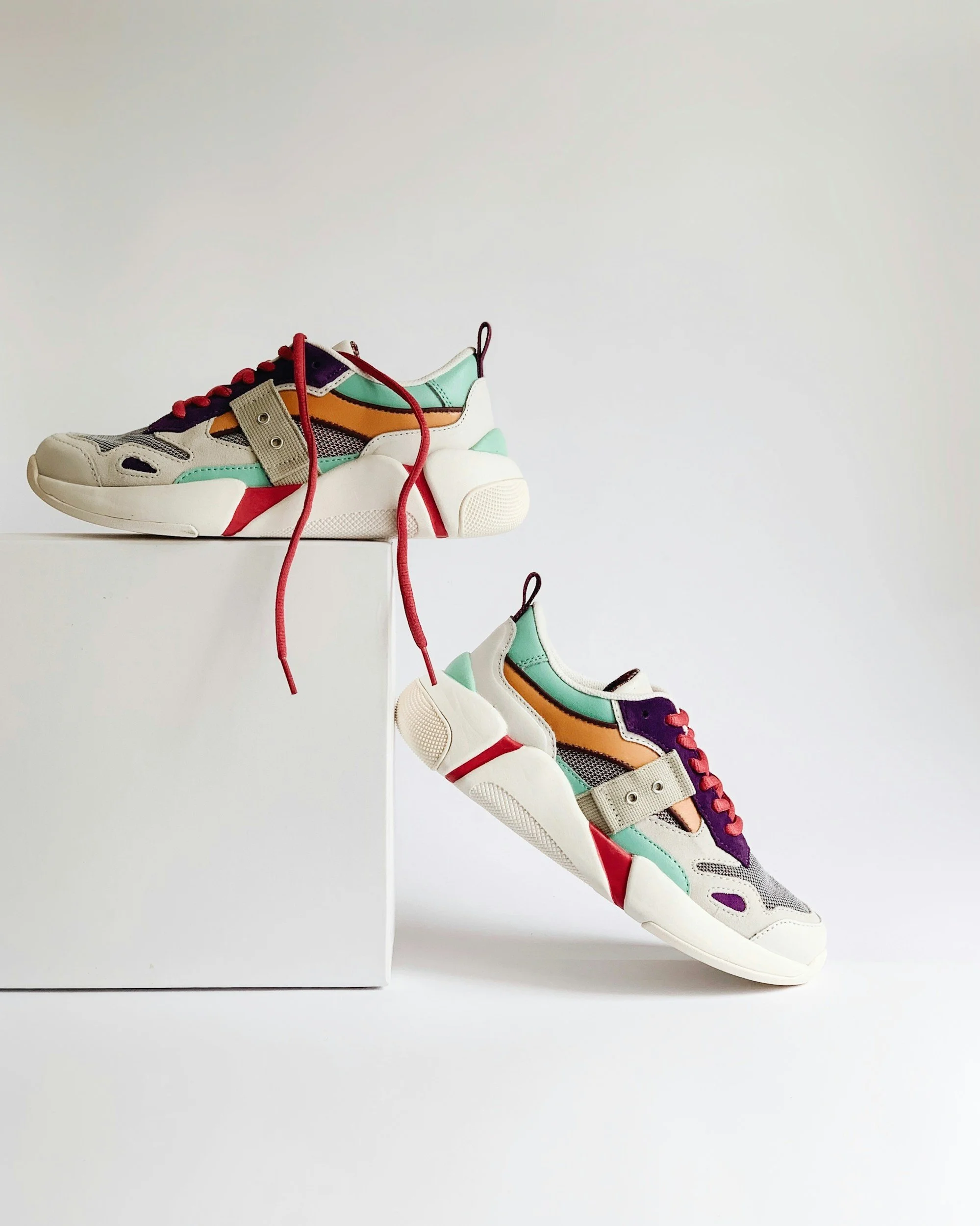 Paio di scarpe sportive multicolori su uno sfondo bianco, con una scarpa appoggiata su una scatola e l'altra sospesa.