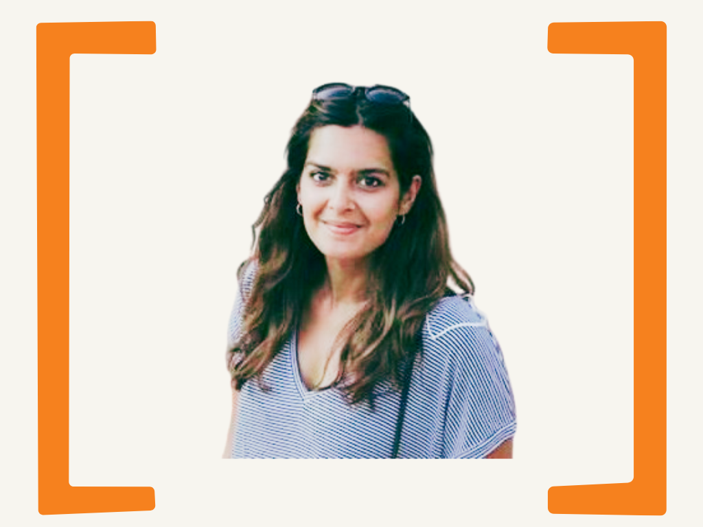 A woman with long, wavy brown hair, wearing sunglasses on her head, a blue and white striped top, and earrings, smiling at the camera, with a minimalistic background and orange framing.