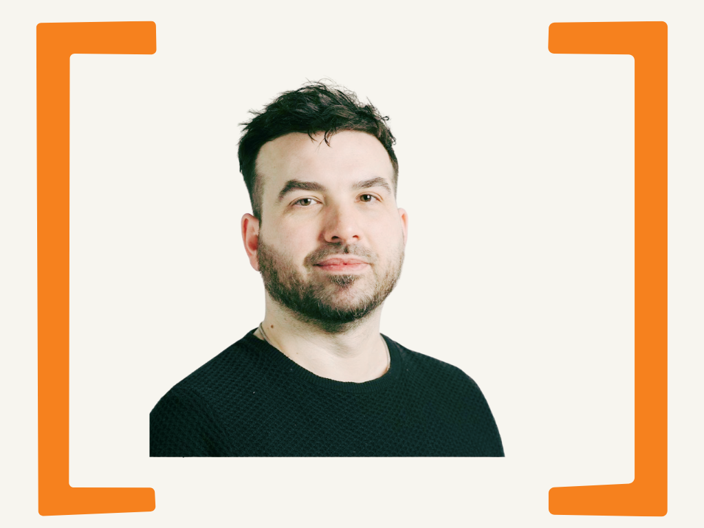 Portrait of a man with dark hair and beard wearing a black shirt, looking at the camera against a plain background, framed by orange brackets.