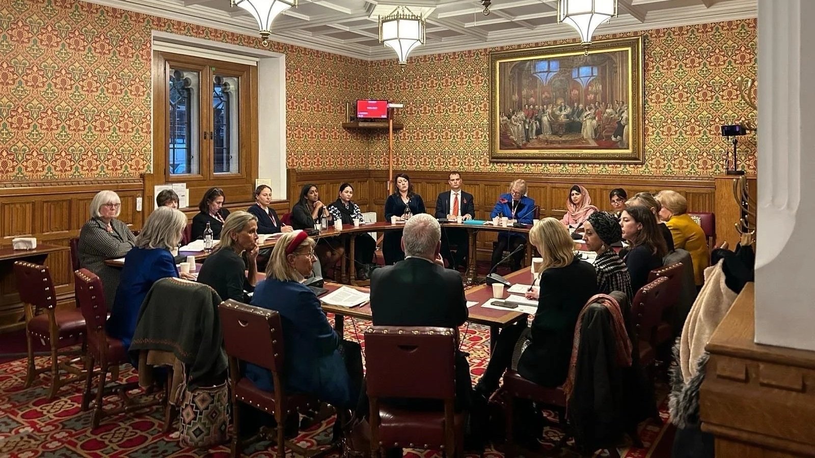 A group of diverse women and men seated around a large, U-shaped conference table in a formal, ornate room with patterned wallpaper, wooden paneling, large windows, and a painting on the wall; some participants are taking notes or listening attentively.