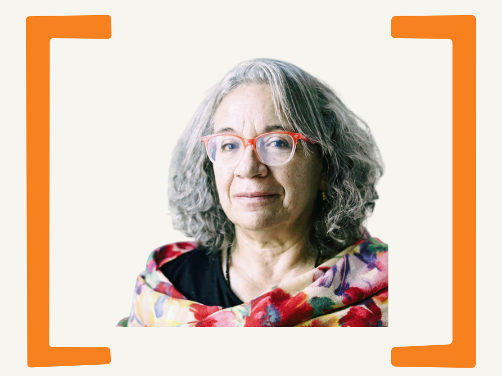 A woman with gray, curly hair and red glasses looks into the camera, wearing a colorful floral scarf over a black top.