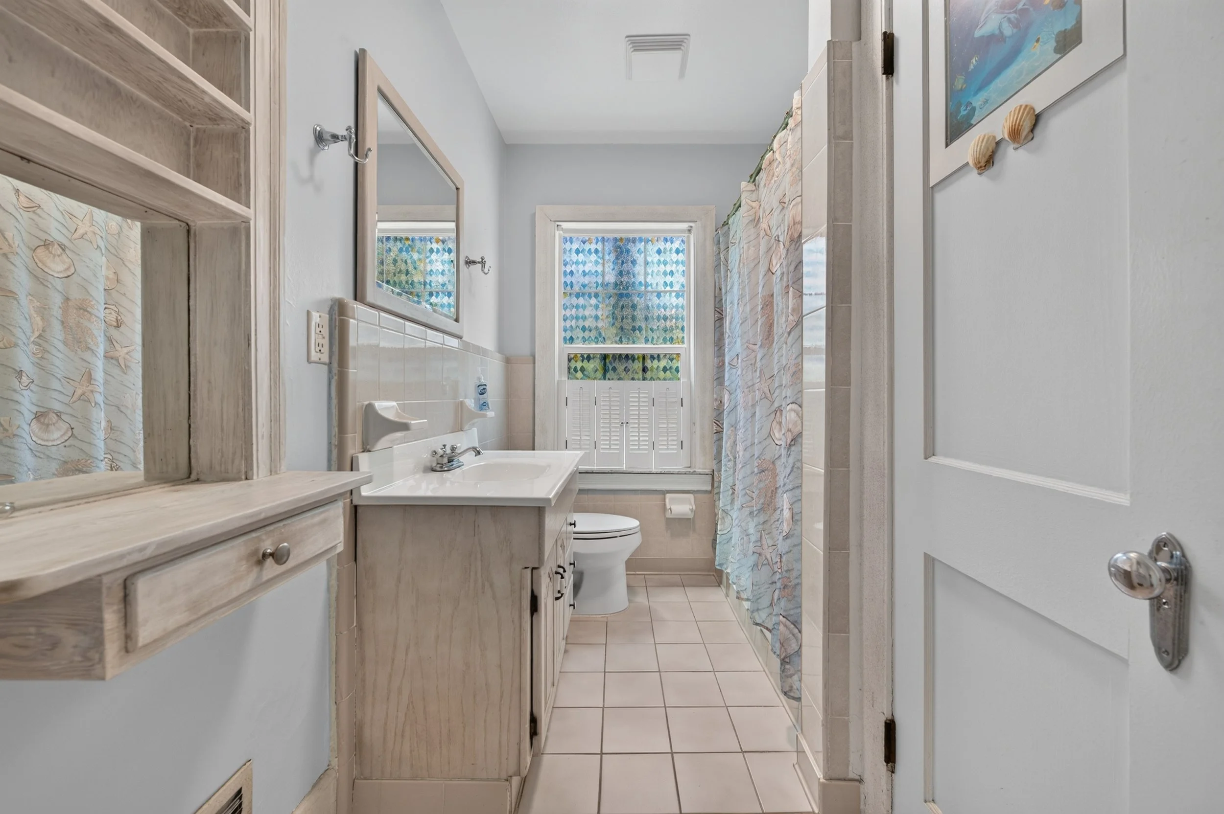 A small bathroom with a white vanity, mirror, toilet, and a shower with sea shell and starfish patterned curtain. The walls are painted light blue, and there are windows with blue and green patterned curtains.
