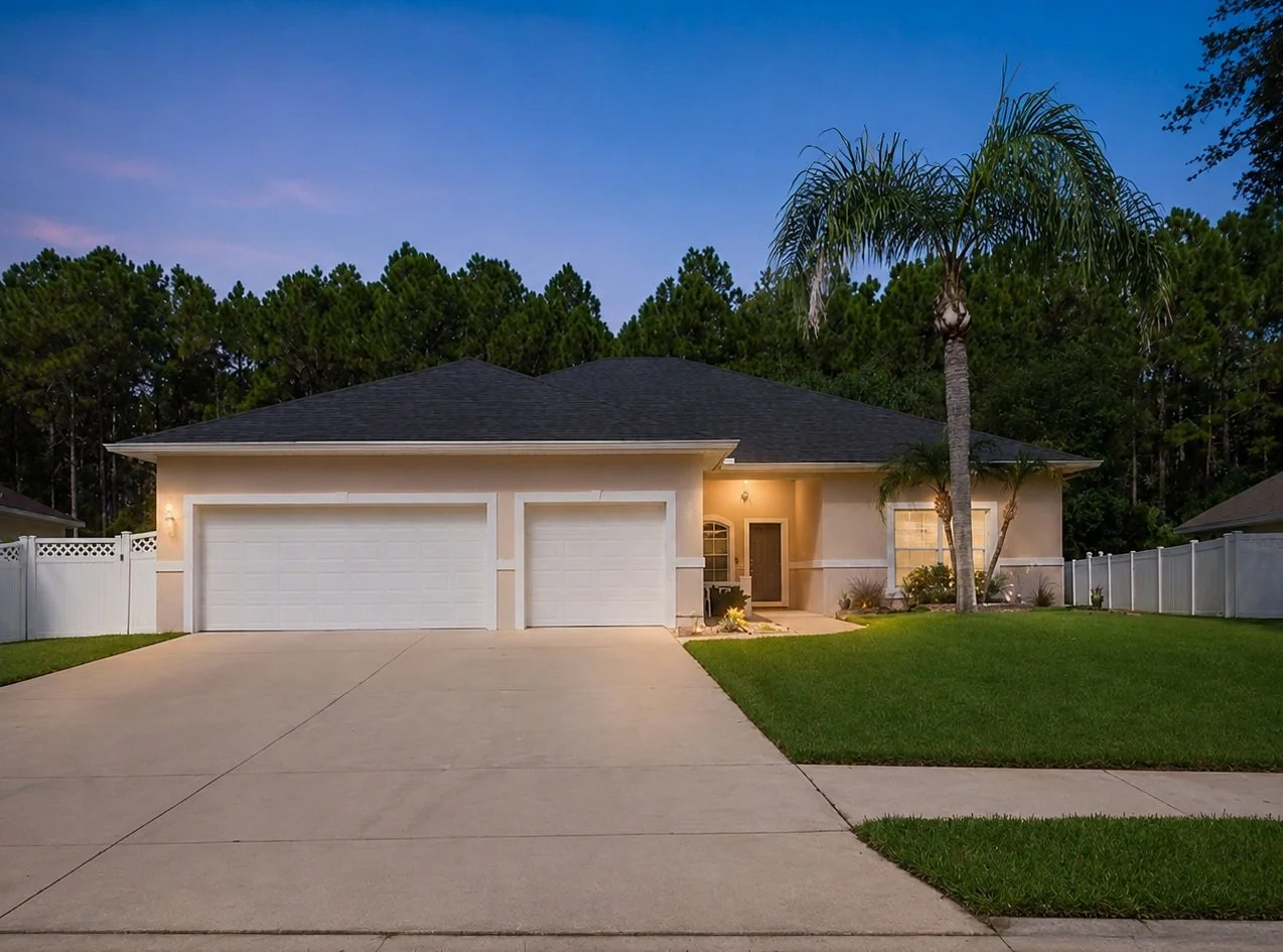 Front view of a suburban house with a three-car garage, palm trees, a well-maintained lawn, and a white fence, during dusk.