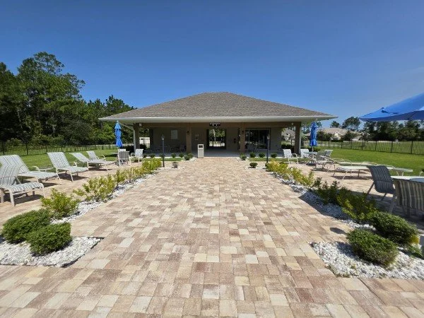 A paved walkway leading to a pavilion with outdoor seating, surrounded by green lawns, trees, and blue umbrellas under a clear blue sky.