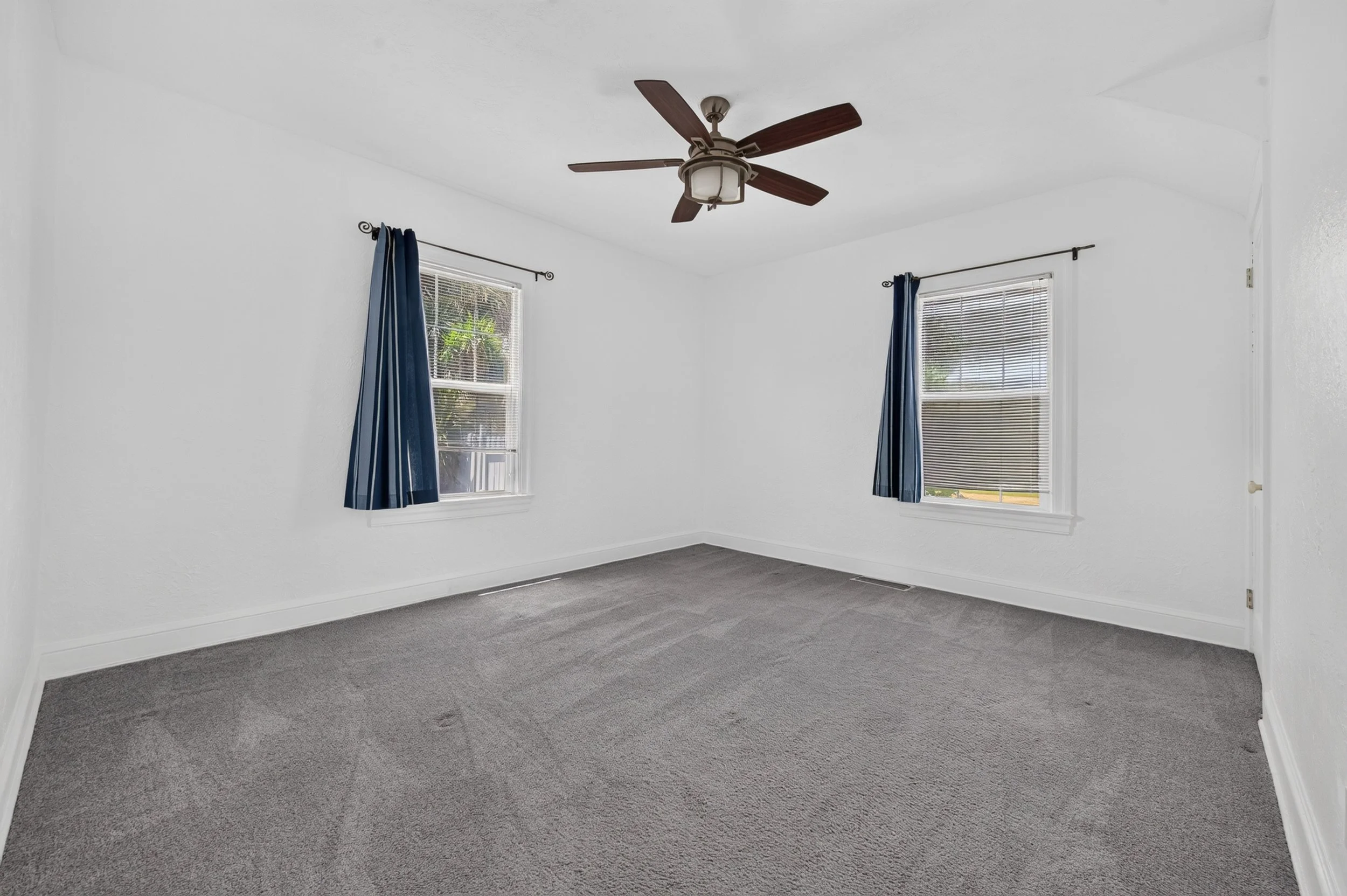 Empty room with white walls, two windows with blinds and blue curtains, a ceiling fan, and gray carpet flooring.
