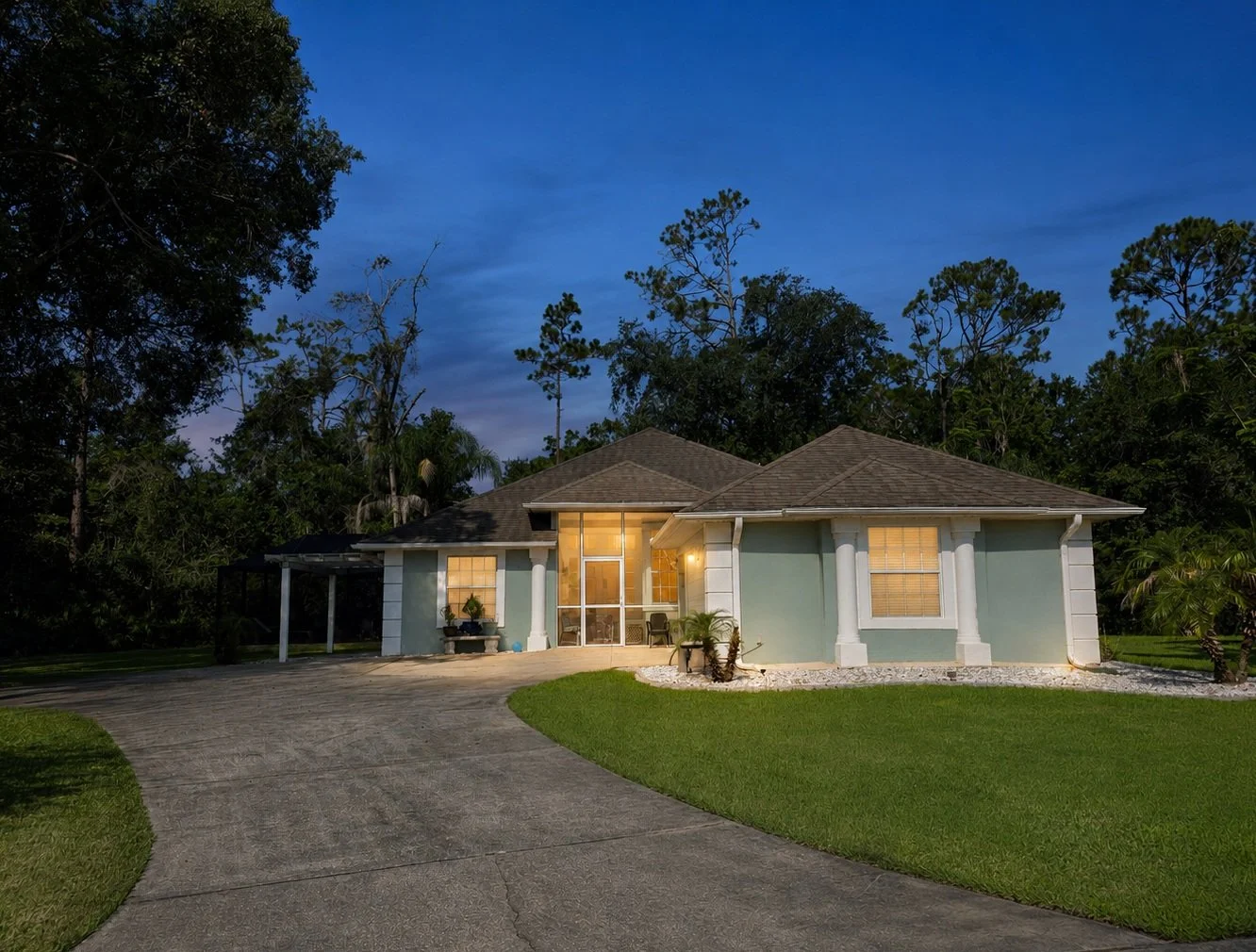 A single-story house with a landscaped yard at dusk, with exterior lighting illuminating the front porch and windows, surrounded by tall trees and a curved driveway.