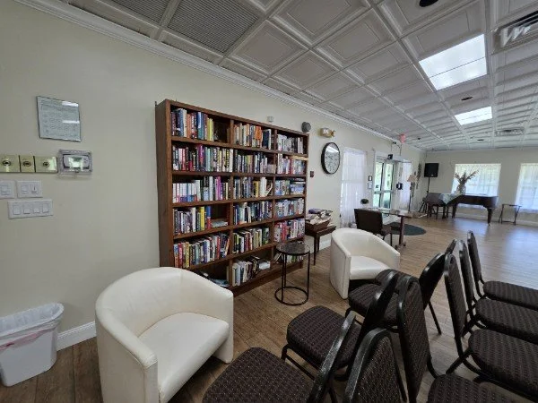A room with a large wooden bookshelf filled with books, surrounded by armchairs and chairs, with a piano in the background and sunlight coming through windows.