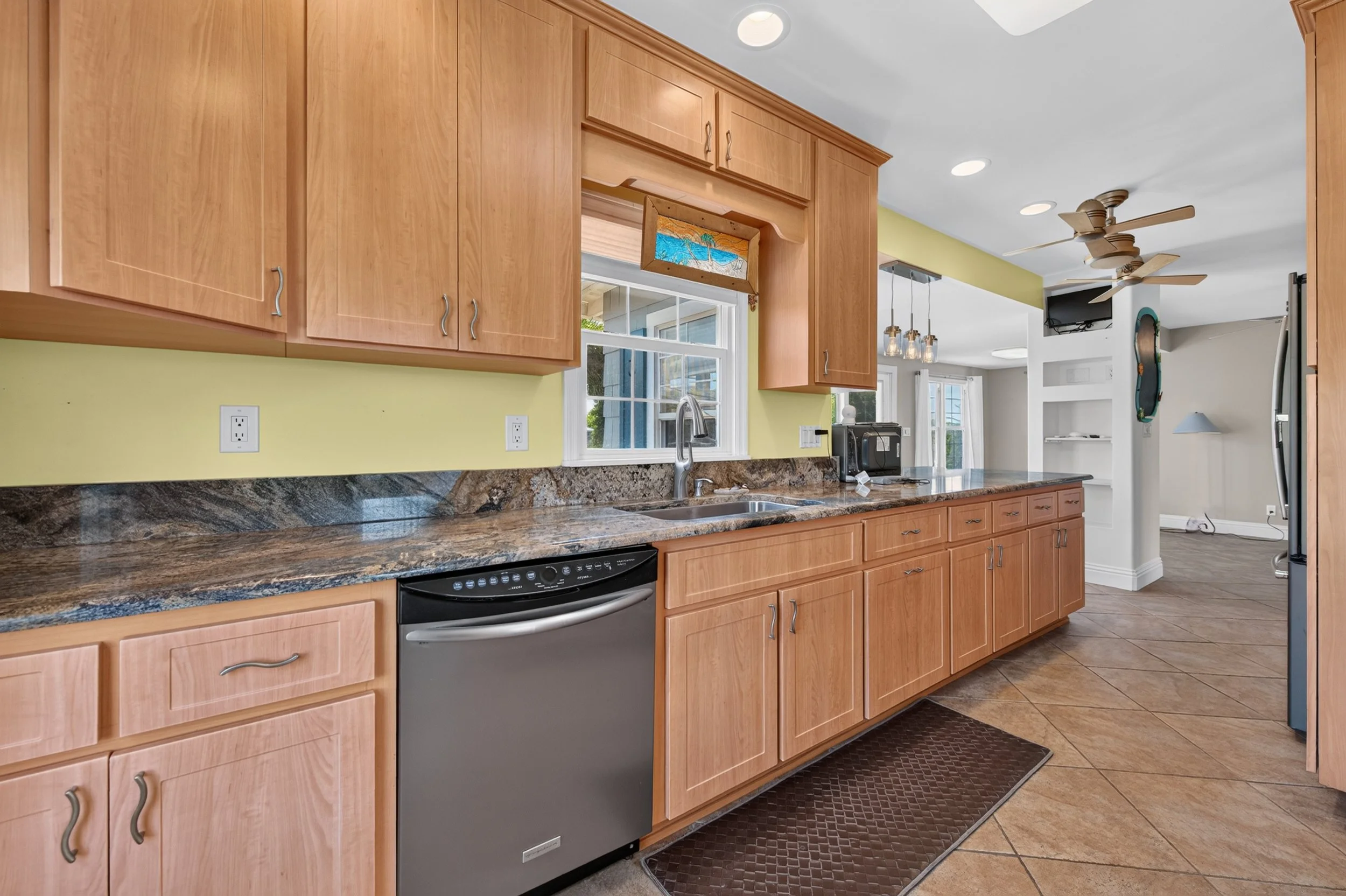 A kitchen with wooden cabinets, granite countertops, a stainless steel dishwasher, a window above the sink, and a partial view of the adjoining living area with a television and ceiling fans.