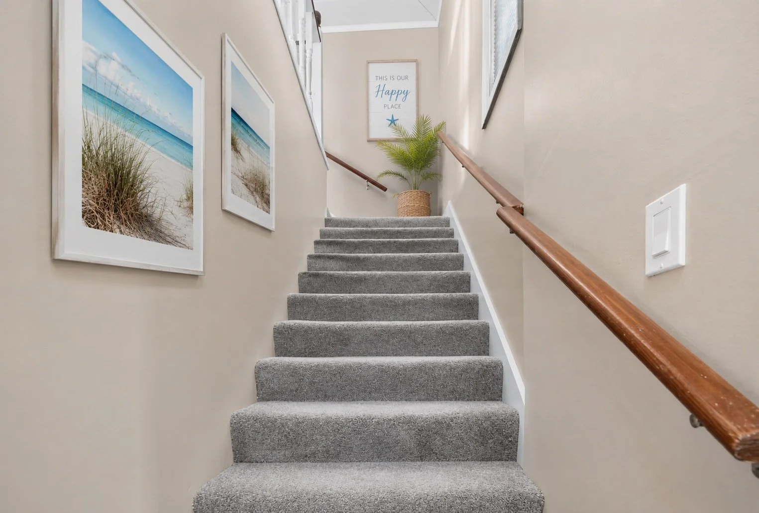 Indoor staircase with gray carpet, beige walls, wooden handrails, wall art of beach scenes, a potted palm plant, and a framed sign that reads 'This is our happy place' at the top of the stairs.