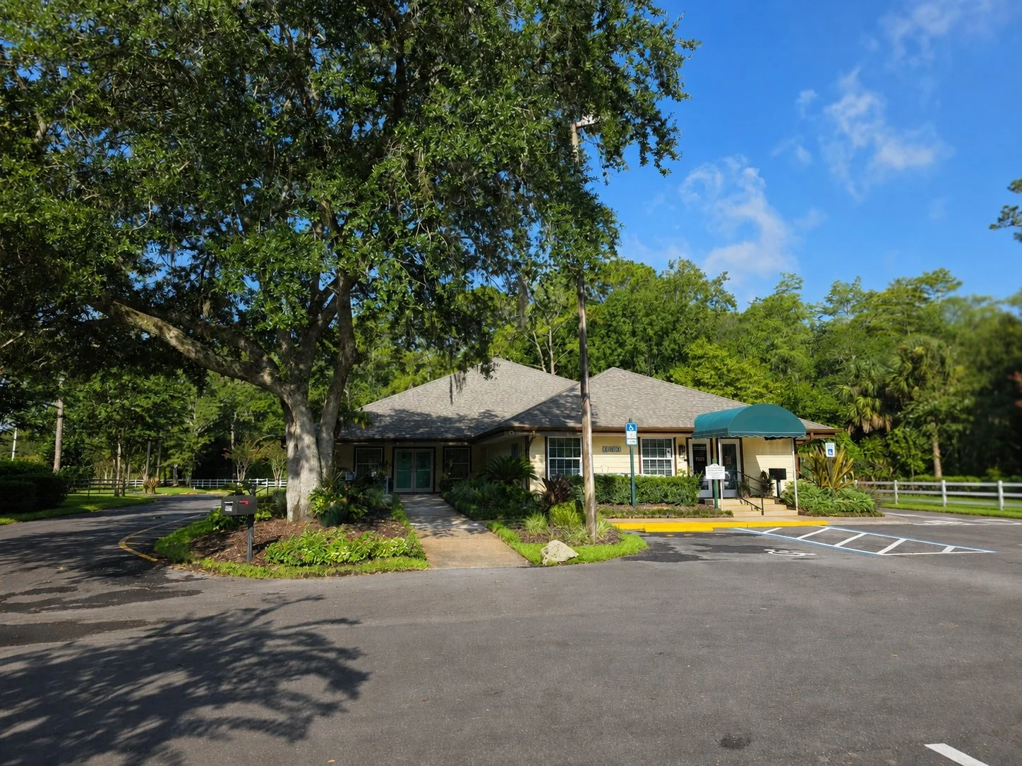 A small, single-story building with a grey roof and beige walls, surrounded by green trees and plants. There are parking spaces in front, including designated handicapped spaces, and a sidewalk leading to the entrance. The sky is clear with a few clo