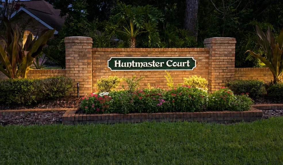 A brick sign with a green nameplate reads 'Huntmaster Court' and is surrounded by well-maintained flowers and greenery.
