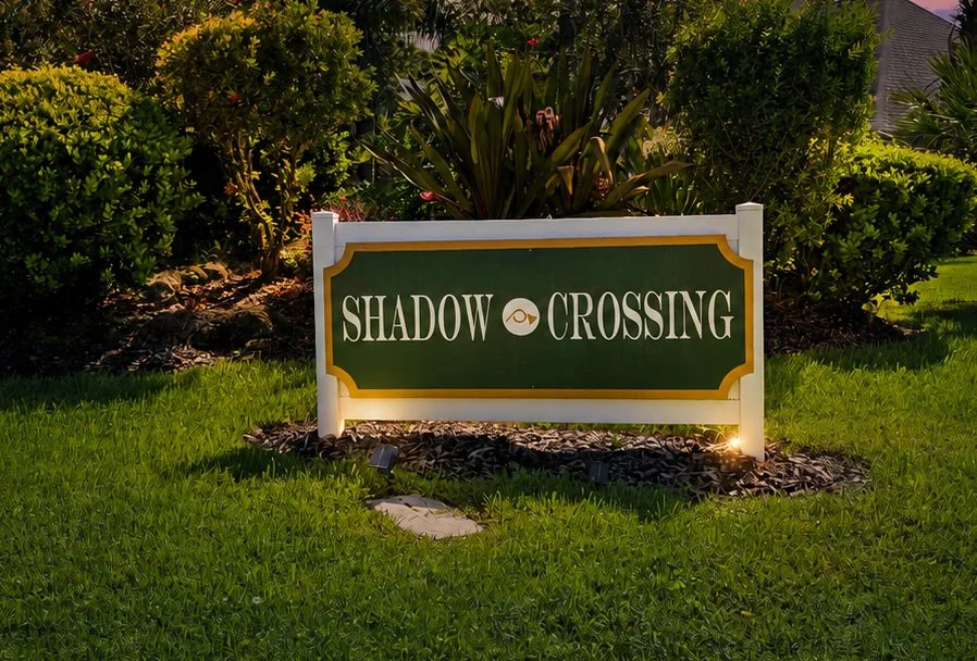 A sign that reads 'Shadow Crossing' with a decorative border, placed on a lush green lawn surrounded by bushes and plants in a landscaped garden.