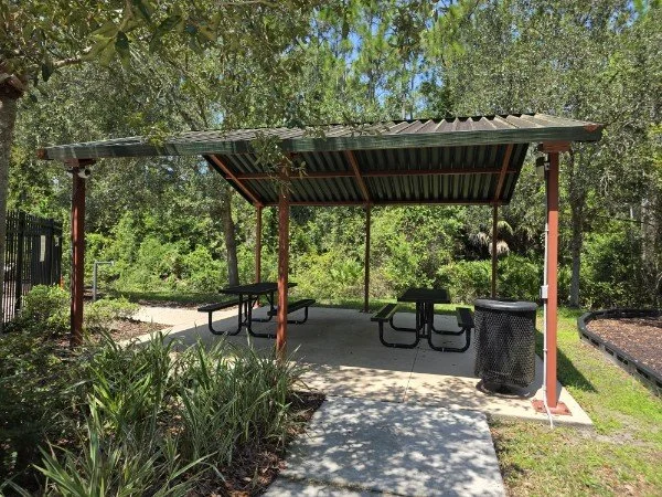 A shaded picnic area with a metal roof, three picnic tables, a trash can, and surrounding greenery.