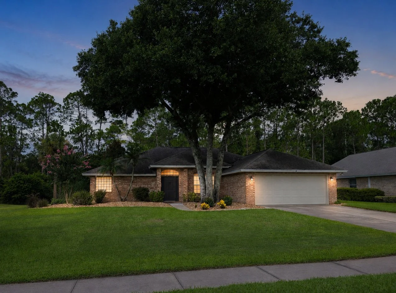 A brick house with an attached garage, front yard, and large tree in a suburban neighborhood during sunset.