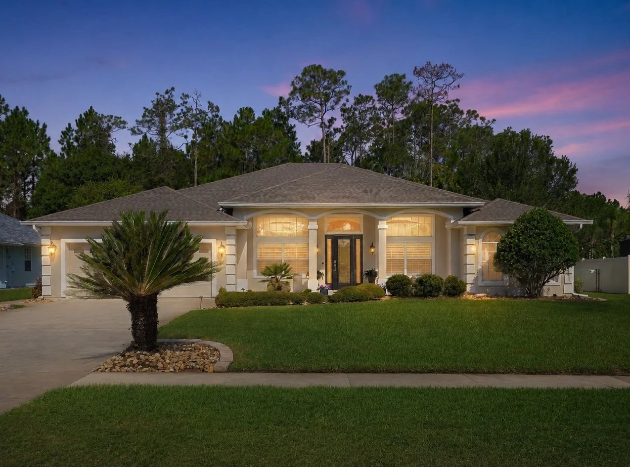 A well-lit suburban house at dusk with a manicured lawn, palm trees, and front porch lights.