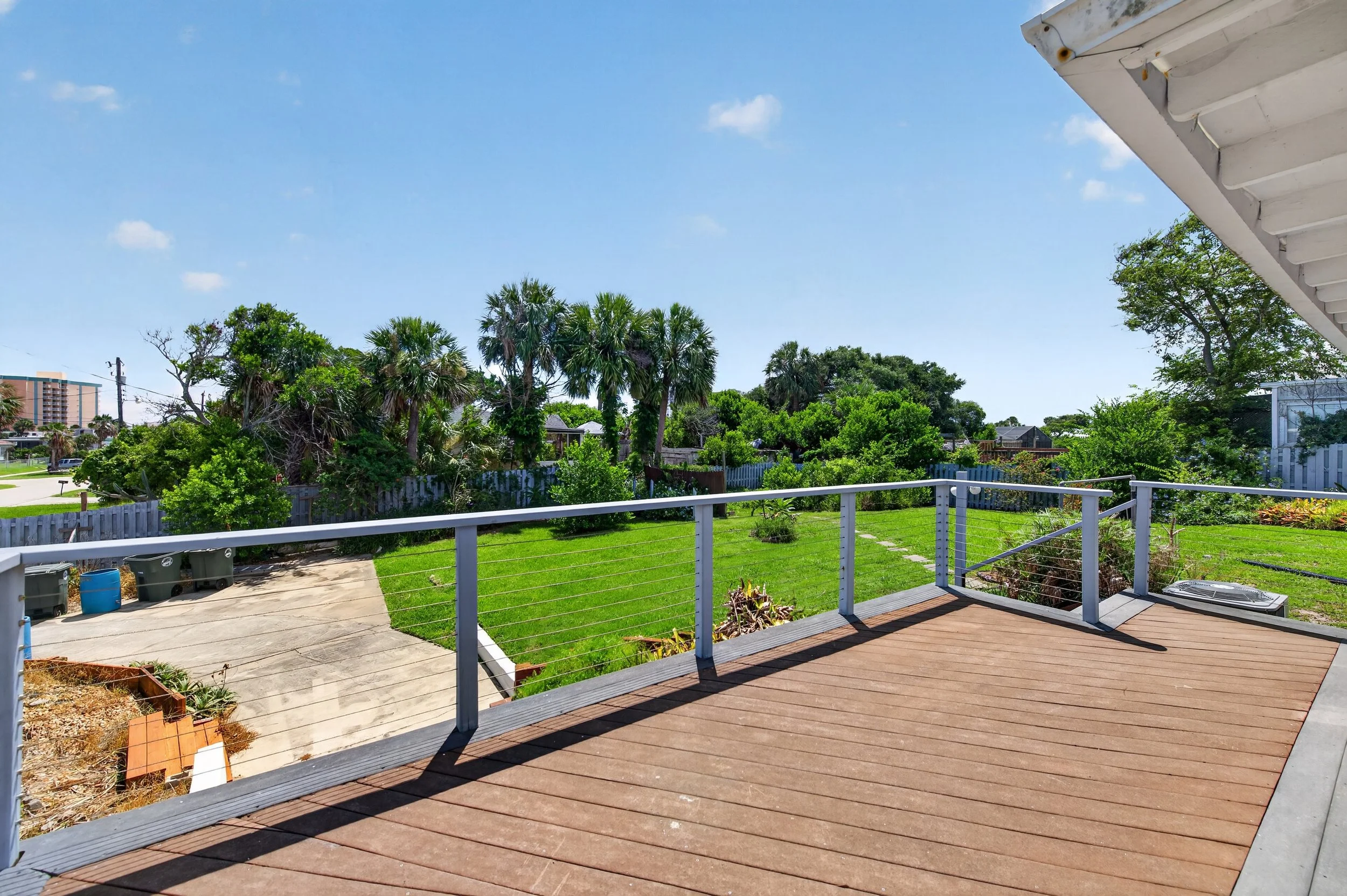View from a porch or deck overlooking a backyard with green grass, shrubs, and tall trees, under a clear blue sky with a few clouds.