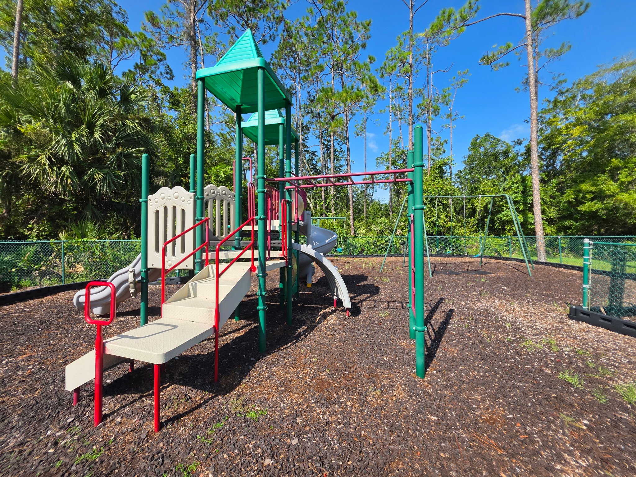 Colorful playground with slides, swings, and climbing structures situated in a forested area with tall trees and a clear blue sky.
