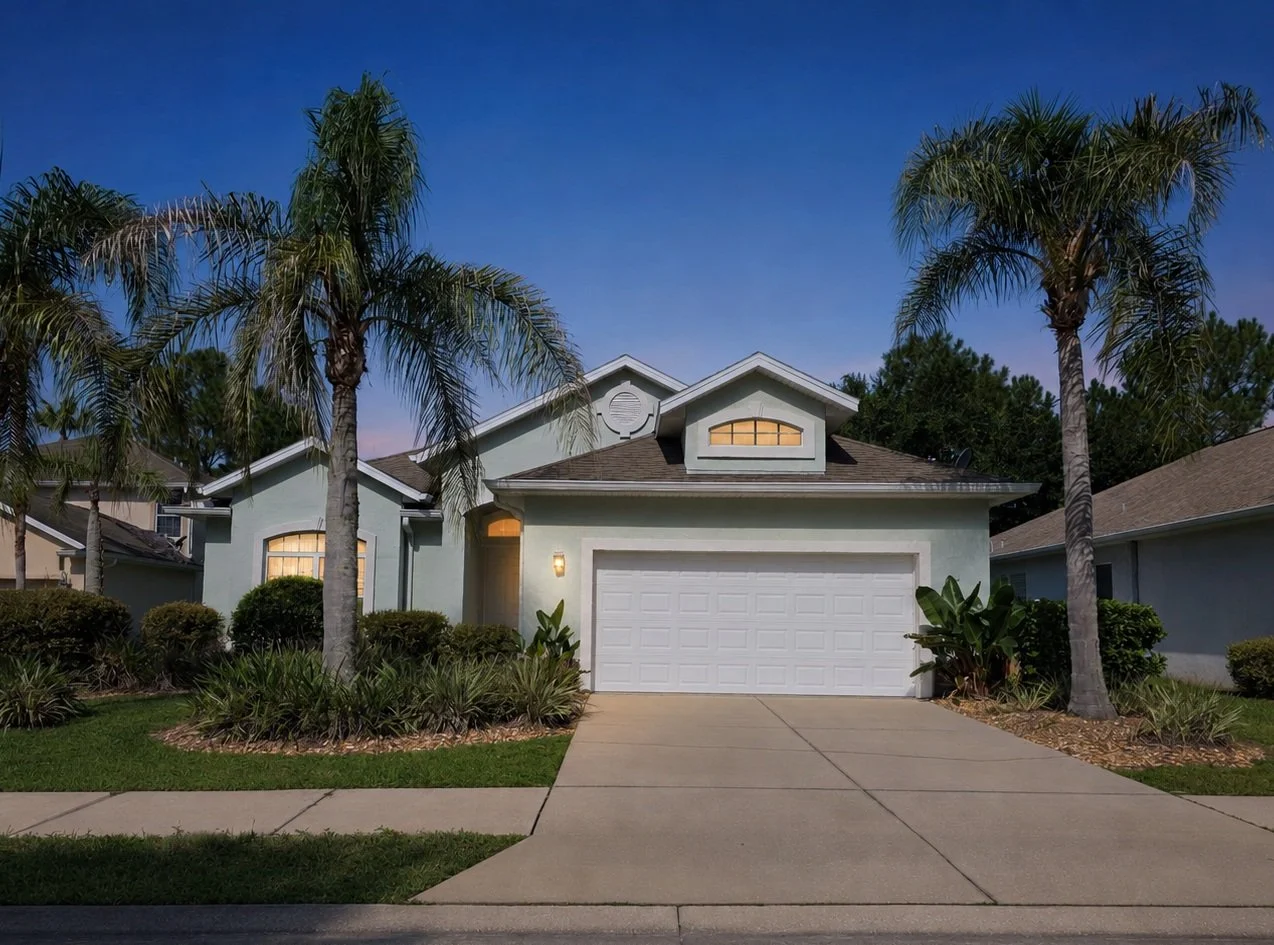 Front view of a suburban house with a two-car garage, palm trees, and landscaped front yard with bushes and plants, under a clear evening sky.
