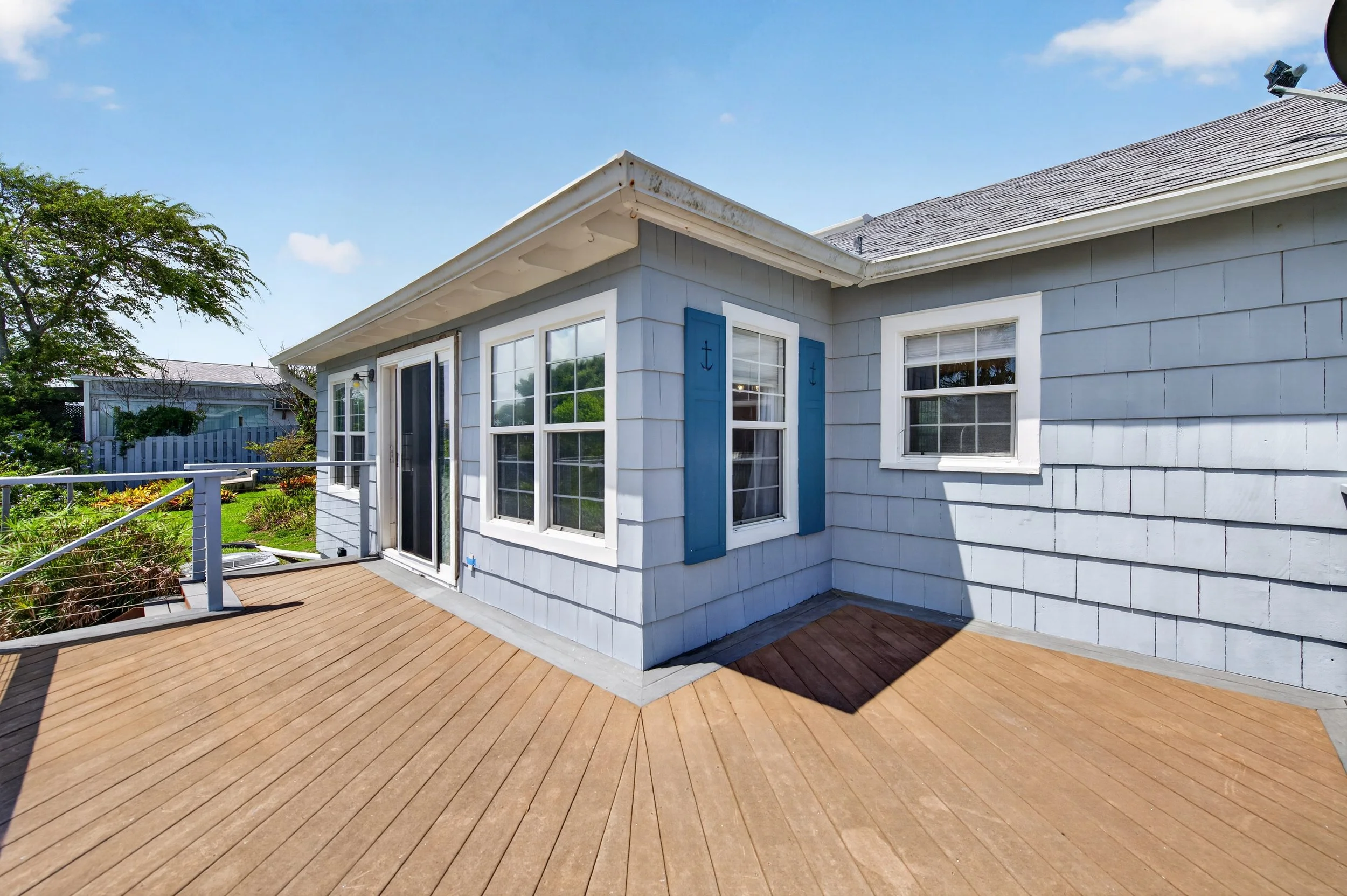 A house with light gray siding, white trim, and blue shutters featuring anchor designs. It has multiple windows and a sliding glass door leading to a wooden deck. The deck surrounds the house and is bordered by a white railing. In the background, the