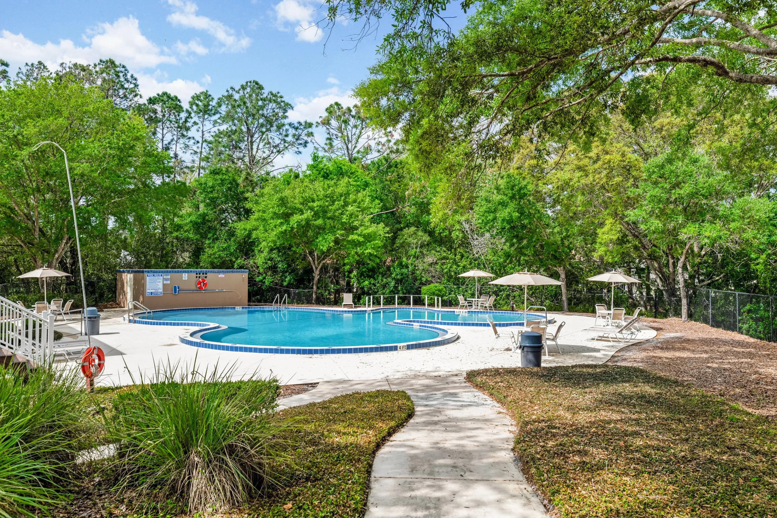 A swimming pool with lounge chairs and umbrellas surrounded by green trees and a bright blue sky.