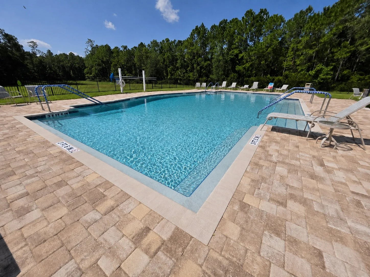 An outdoor swimming pool with clear blue water, surrounded by a brick paver deck, white lounge chairs, and a green tree line in the background under a partly cloudy sky.