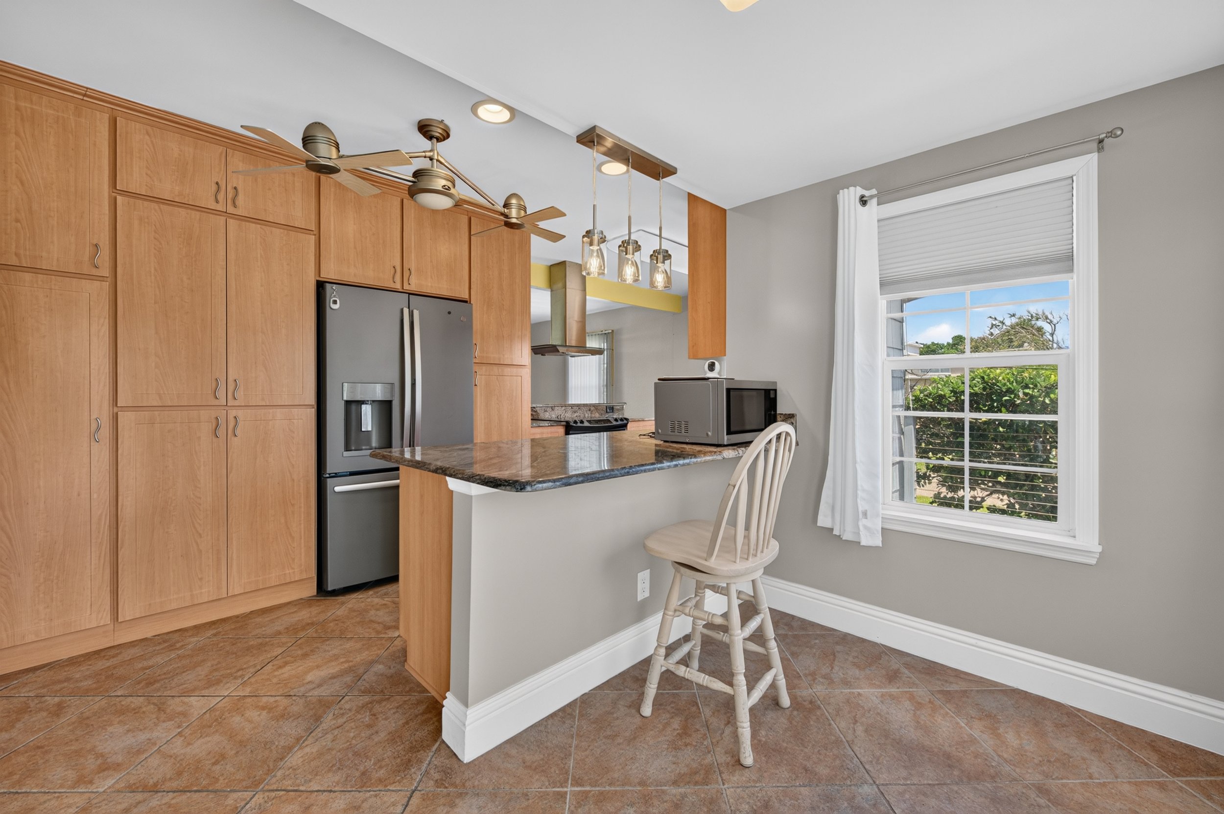Kitchen with wooden cabinets, stainless steel refrigerator, granite countertop, small microwave, window with white curtains, and a bar stool.