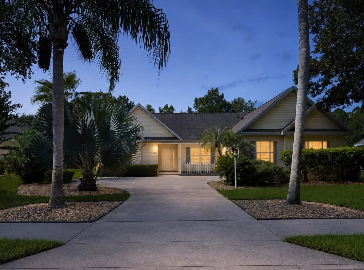 A suburban house at dusk, with a driveway leading to the front door, surrounded by palm trees and greenery.