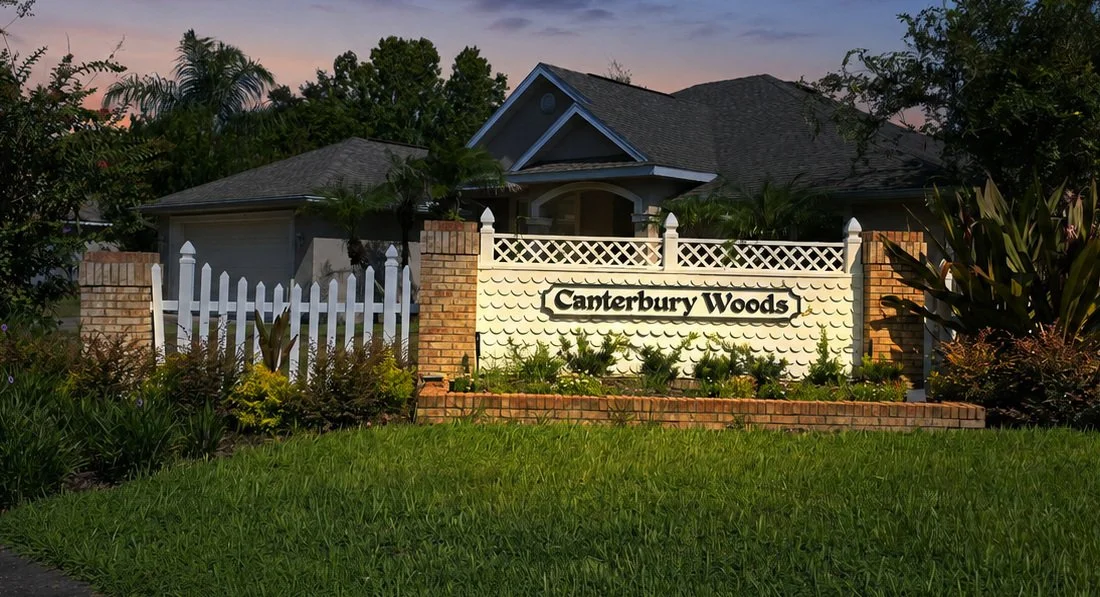 A residential entrance sign for Canterbury Woods, with a white fence, brick and white decorative wall, and lush green plants in front, under a evening sky.