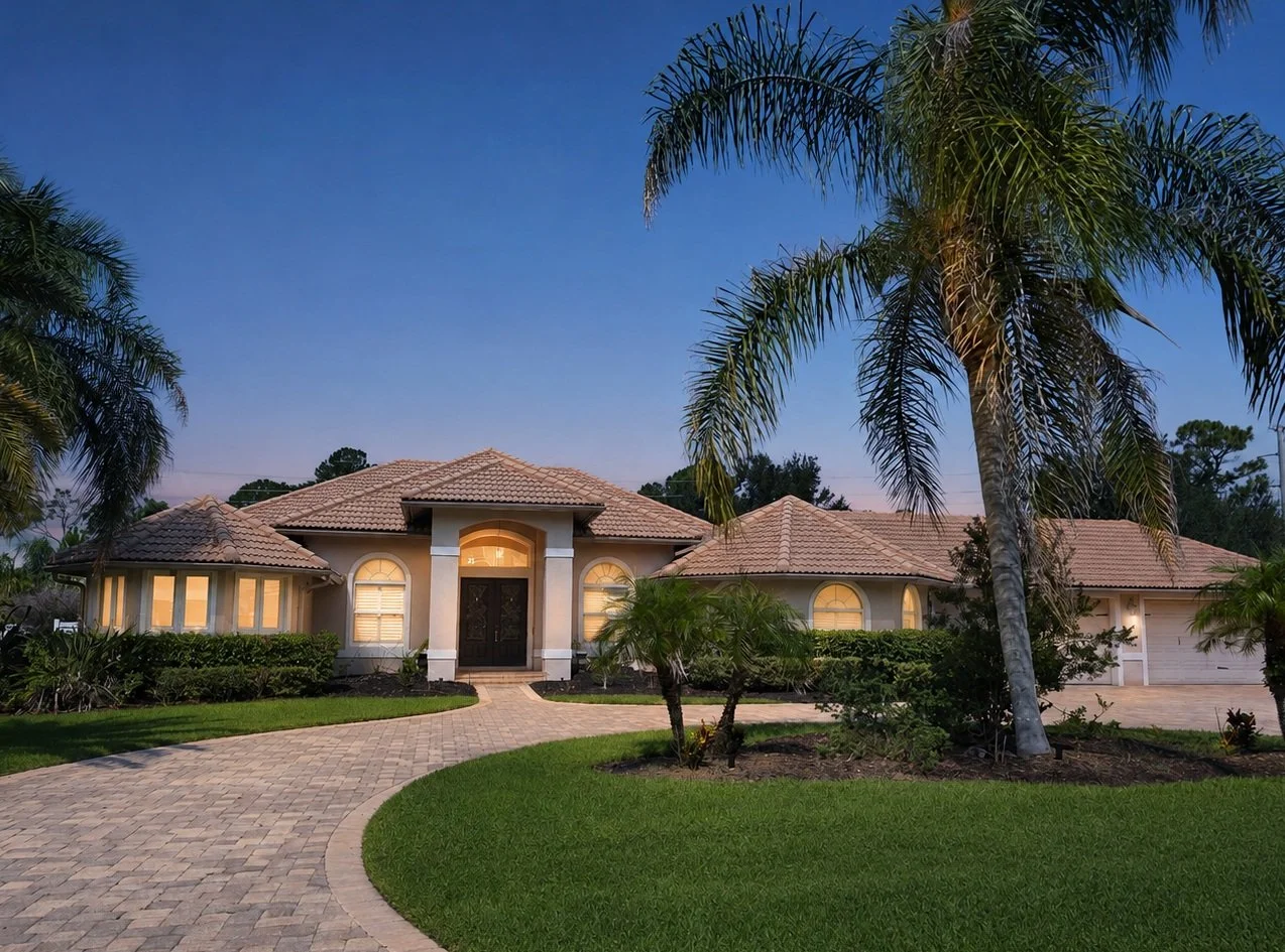 Front view of a large, modern house with a tiled roof, arched windows, and a driveway, surrounded by lush green grass and palm trees at dusk.