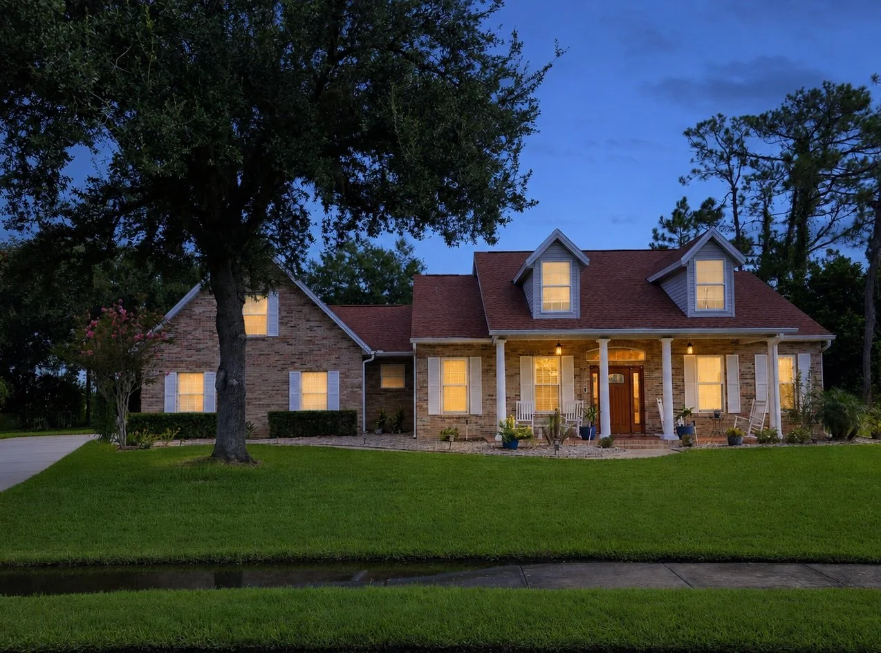 A well-lit brick house with a front porch, white columns, and potted plants, surrounded by lush green grass and trees at dusk.