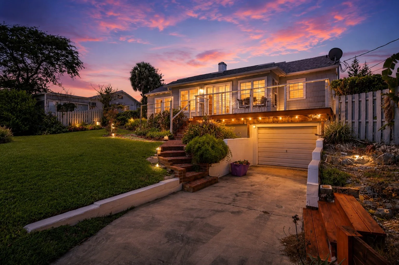 A house with lit windows and a balcony, surrounded by a well-maintained lawn and garden, at dusk with a colorful sky.