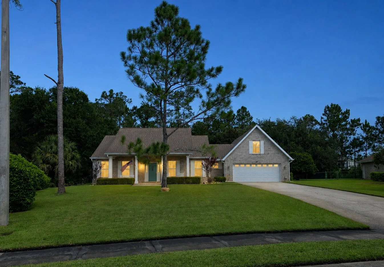 A suburban house with a well-maintained lawn and trees, illuminated windows, a two-car garage, and a driveway during dusk.