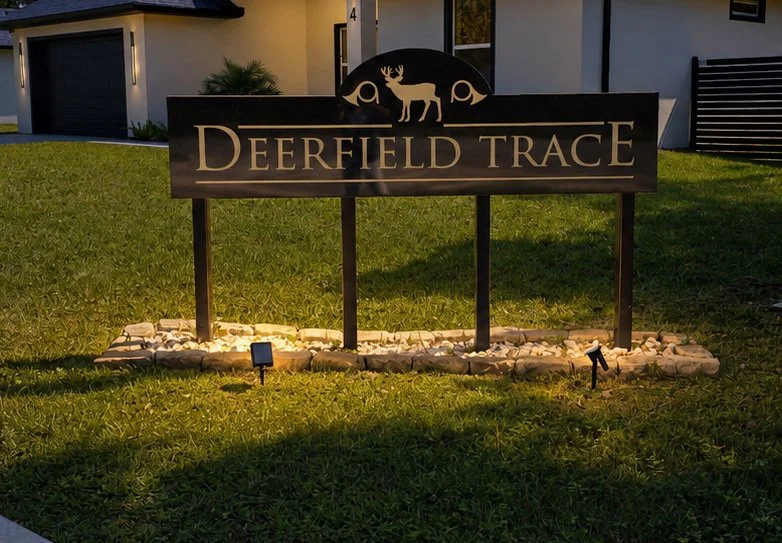 A sign reads "Deerfield Trace" with a silhouette of a deer and two trees above the text, illuminated at night in a yard with grass and a house in the background.