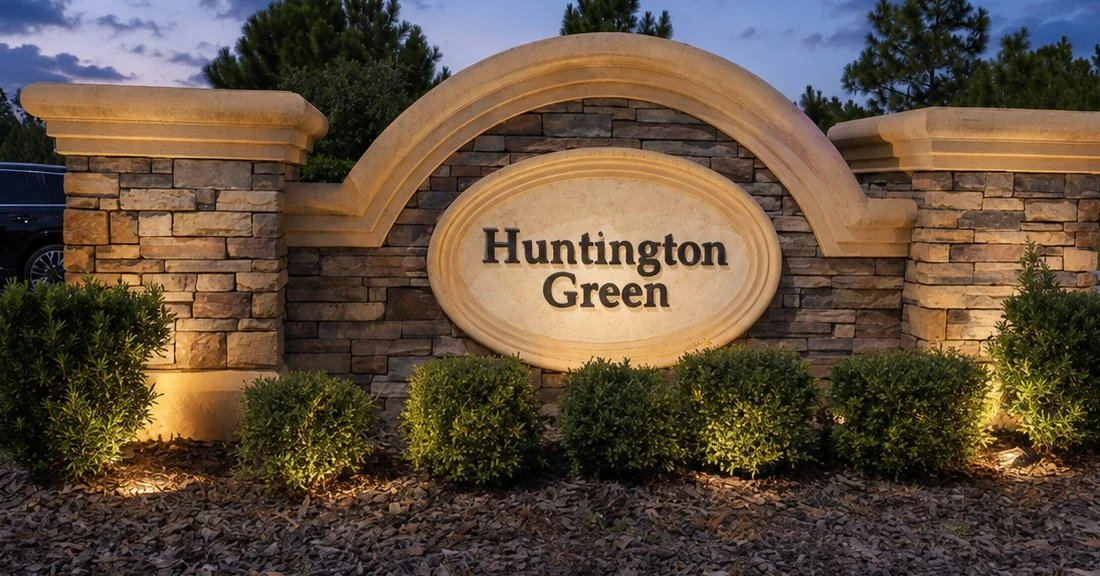 A stone sign reads 'Huntington Green' in front of a landscaped area with bushes and trees, illuminated by outdoor lights during dusk.