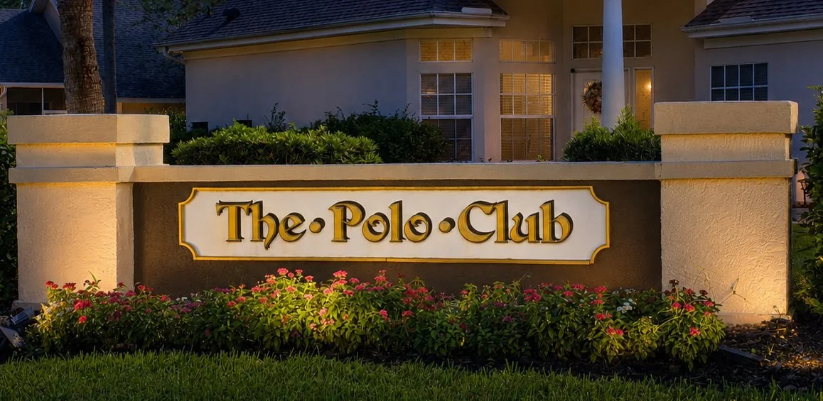 Sign for The Polo Club at the entrance of a residential community, with flowers and building in the background.