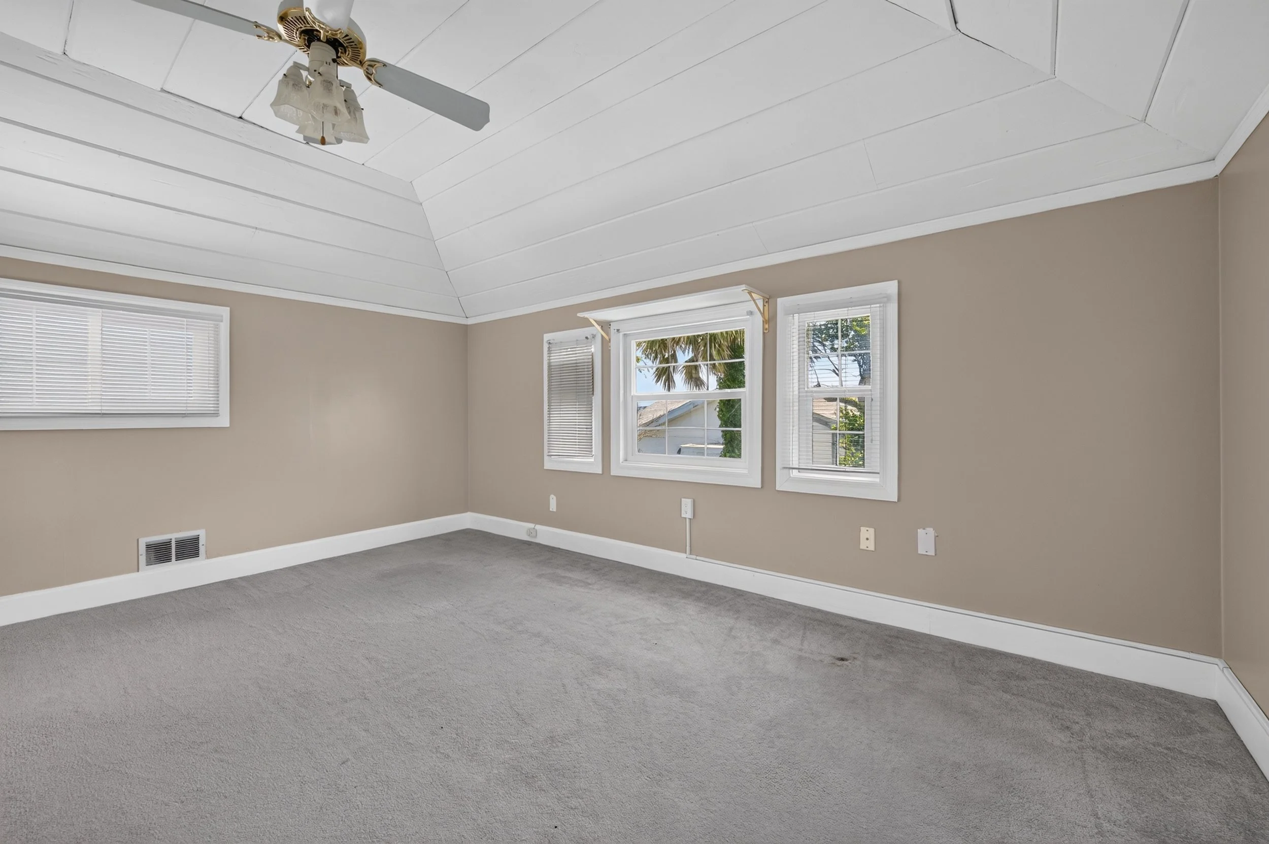 A bright empty room with beige walls, white trim, three windows with blinds, a ceiling fan, and gray carpet flooring.