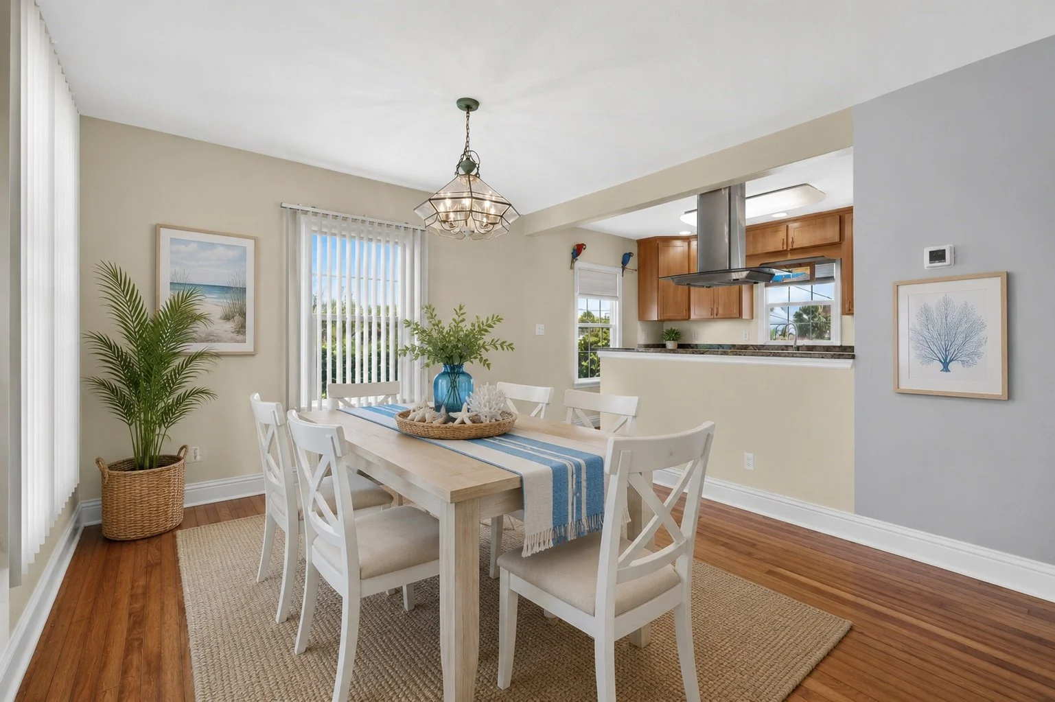 Dining room with white table and chairs, blue and white table runner, globe plant, wall art, and view into kitchen with wooden cabinets and window