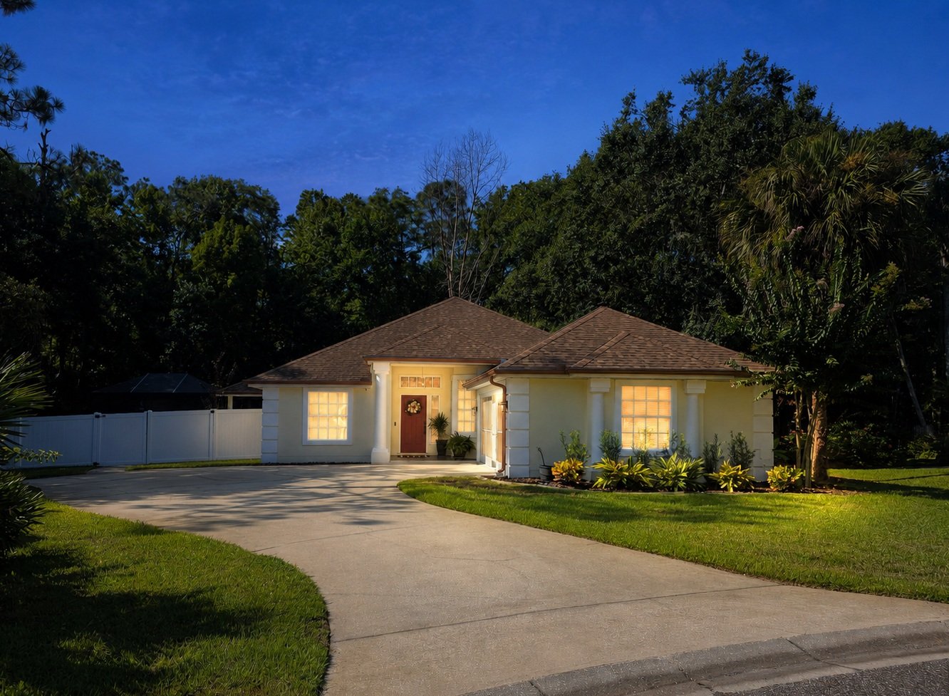 A well-lit, single-story house with white exterior walls, a brown roof, and illuminated windows at dusk. The house has a curved driveway, a small porch, and a garden with shrubs and flowering plants.